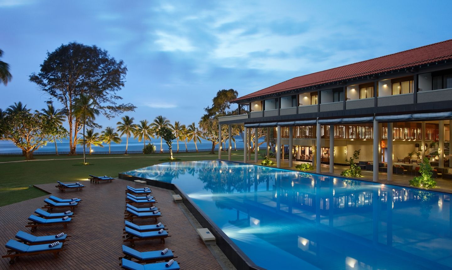 نمای باز از استخر بزرگ هتل ساحلی لوکس در هنگام عصر با تخت‌های آفتاب‌گیر آبی رنگ و درختان نخل در پس‌زمینه.