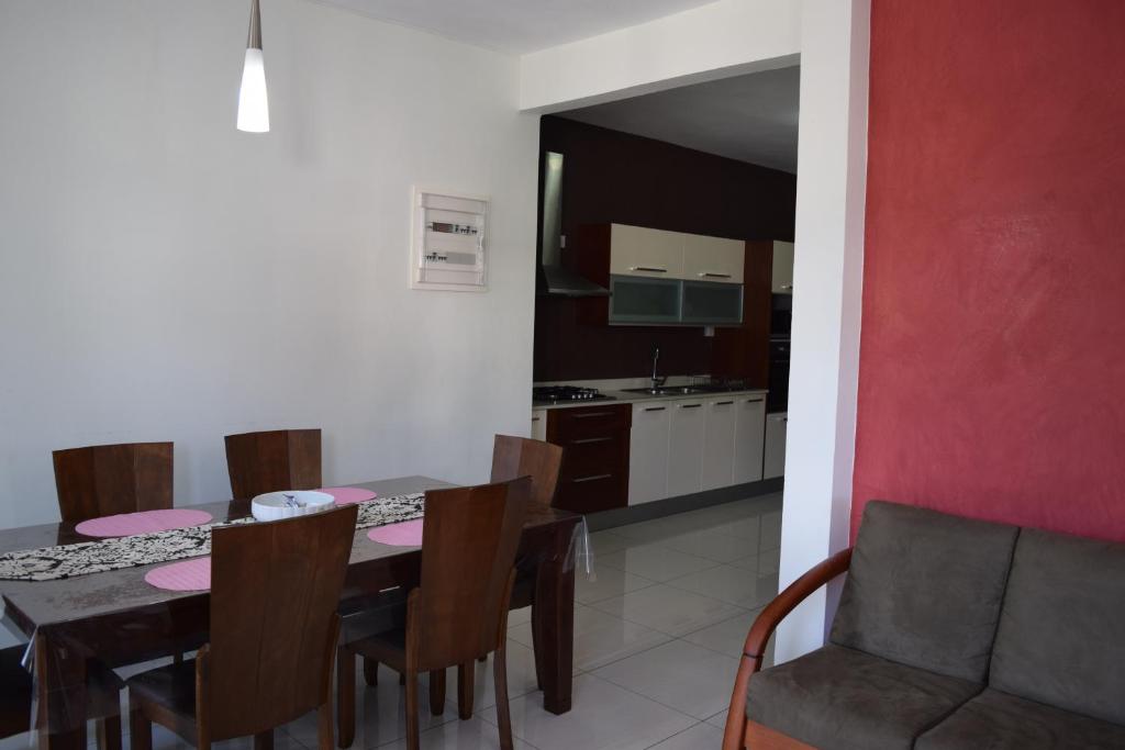 نمای داخلی یک آپارتمان مدرن با میز ناهارخوری چوبی شش نفره، صندلی‌های قهوه‌ای و آشپزخانه مجهز در پس‌زمینه با دیوارهای سفید و قرمز.