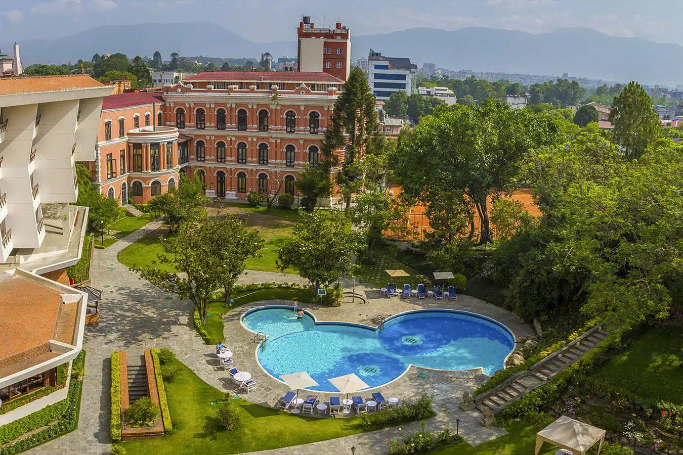 نمای هوایی از حیاط هتل پنجستاره یاک و یتی کاتماندو، شامل استخر شنای فضای باز با طراحی خاص، فضای سبز، زمین تنیس و ساختمان تاریخی قصر مانند با معماری کلاسیک نپالی.